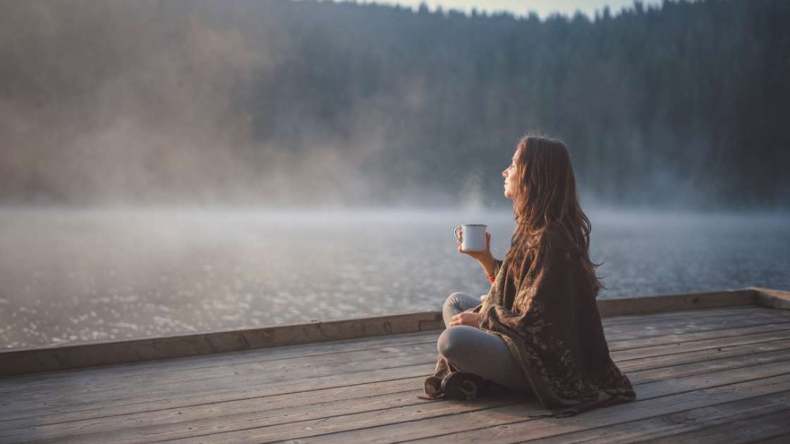 Um ihren Stres abzubauen, sitzt eine Frau auf einem Steg am See und genießt achtsam eine Tasse Tee.